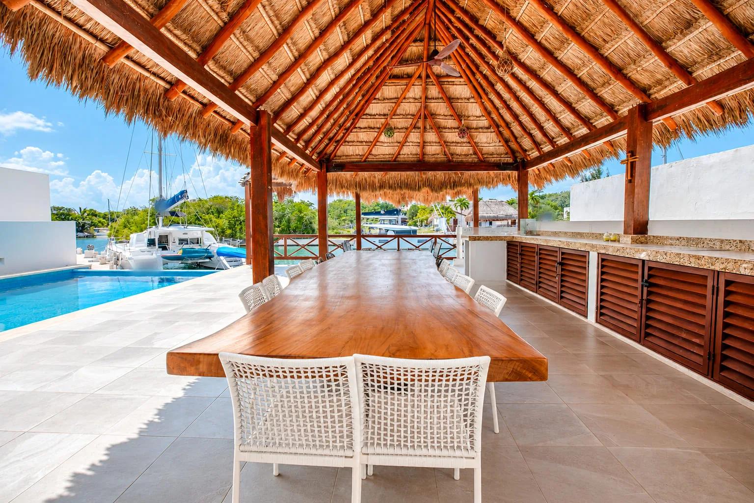 Palapa with a long wooden dining table, white chairs and a bar counter with cabinets beside the pool, with a catamaran docked on the canal at Casa Playa Cataluña, Puerto Aventuras