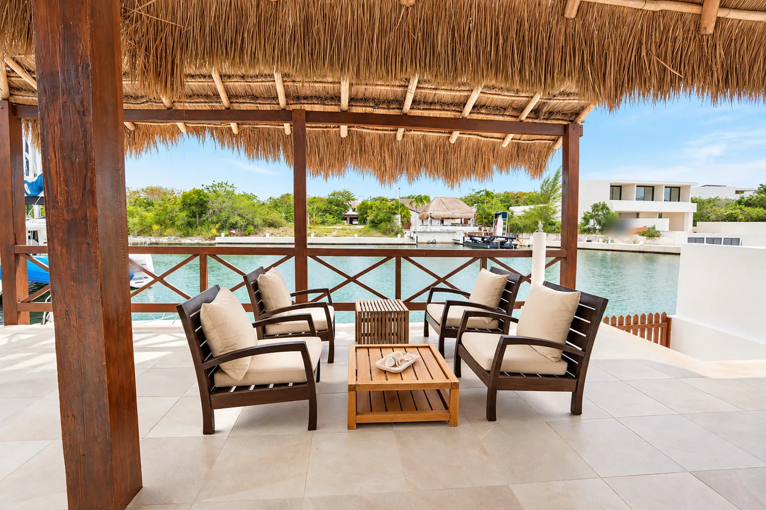 Terrace under a thatched palapa with four wooden armchairs and a coffee table, wooden railing and a view of the canal and houses on the opposite bank at Casa Playa Cataluña, Puerto Aventuras