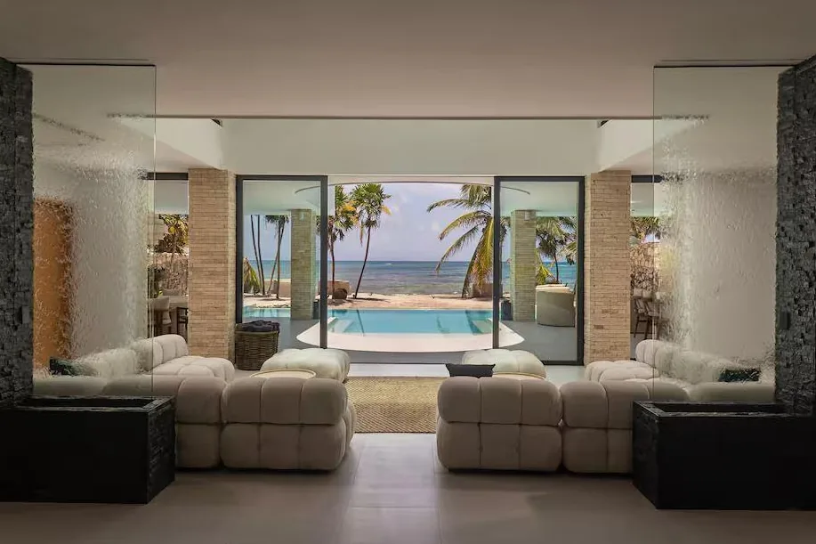 Sala con muros de agua y salida a la alberca con vista al mar entre palmeras, en Palacio del Mar